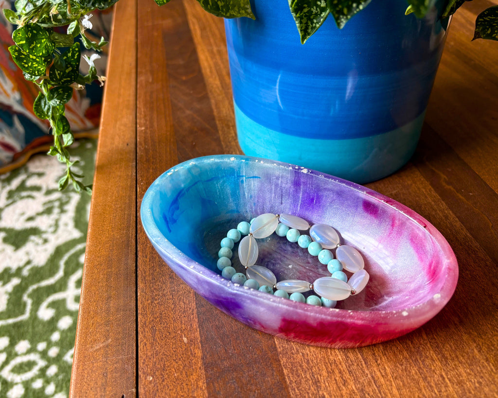 A handmade resin tray with a blue, purple, and pink watercolor design, holding a light blue beaded bracelet with white oval beads, set on a wooden table next to a blue ceramic pot.