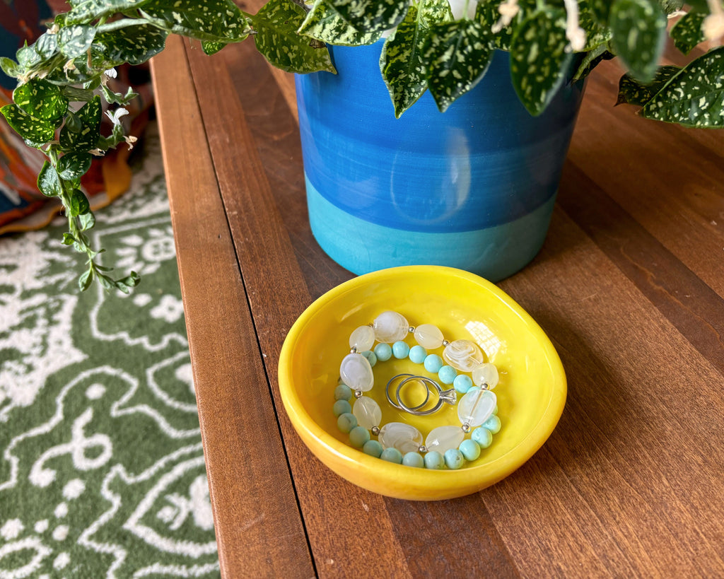 A yellow resin jewelry tray holding a beaded bracelet and rings, placed on a wooden table next to a blue and green potted plant.