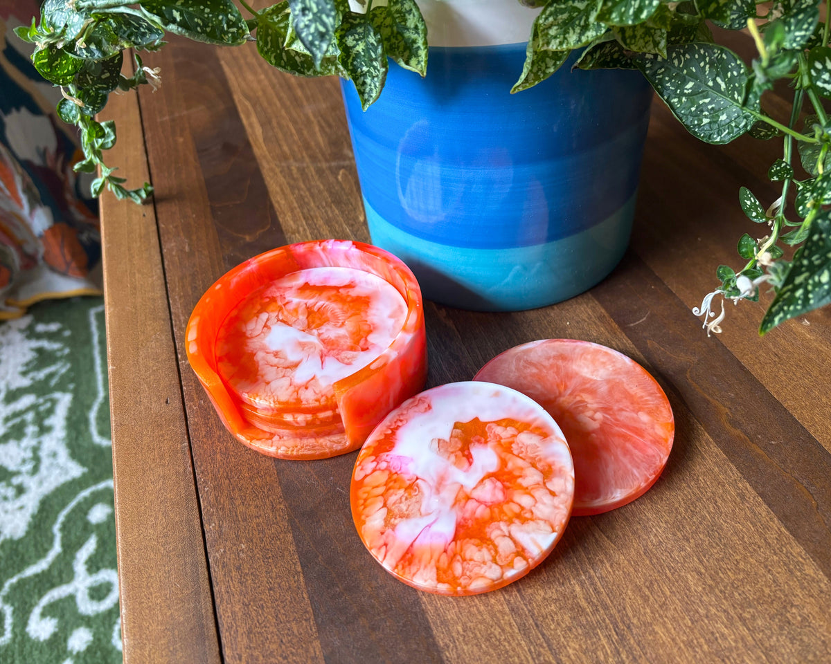 Round coasters with a white and orange marbled pattern, with one standing upright to show its small pouring spout, displayed on a wooden table next to a blue ceramic pot with green trailing plants.