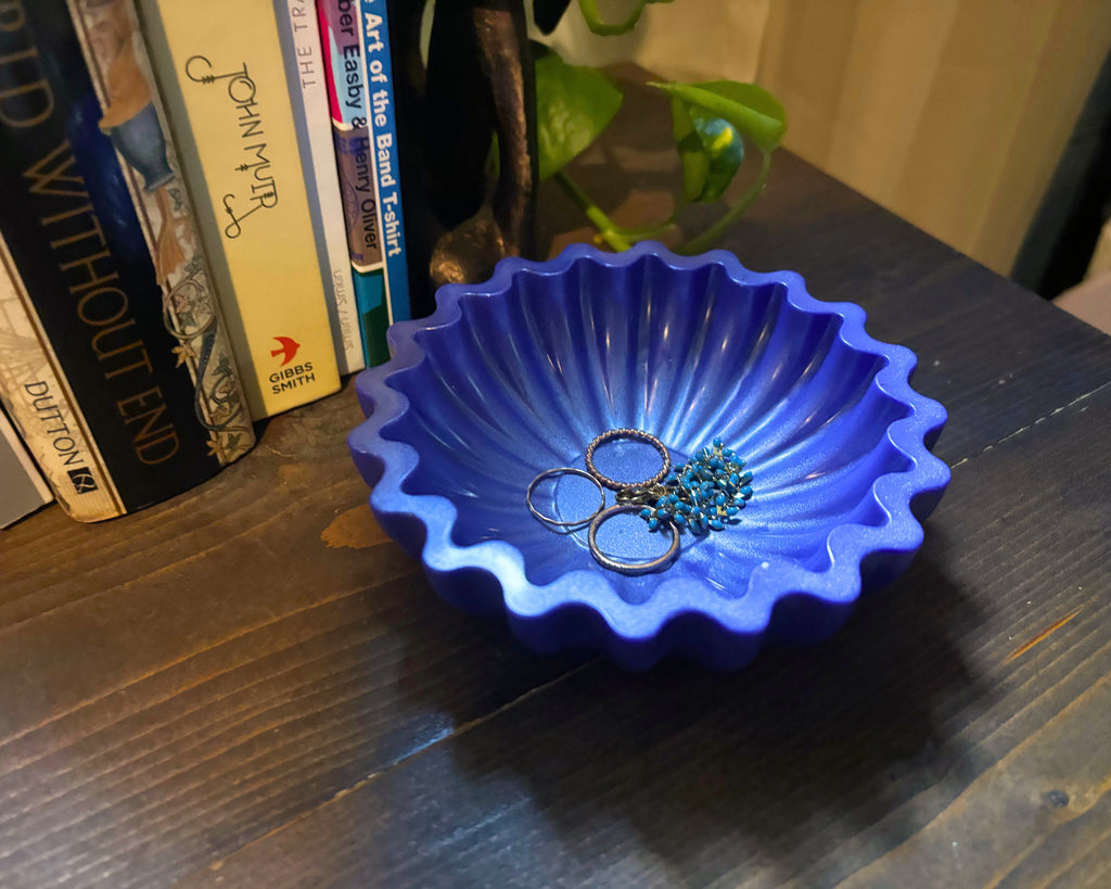 A periwinkle scalloped resin jewelry organizer bowl containing several rings, placed on a dark wooden table next to books.