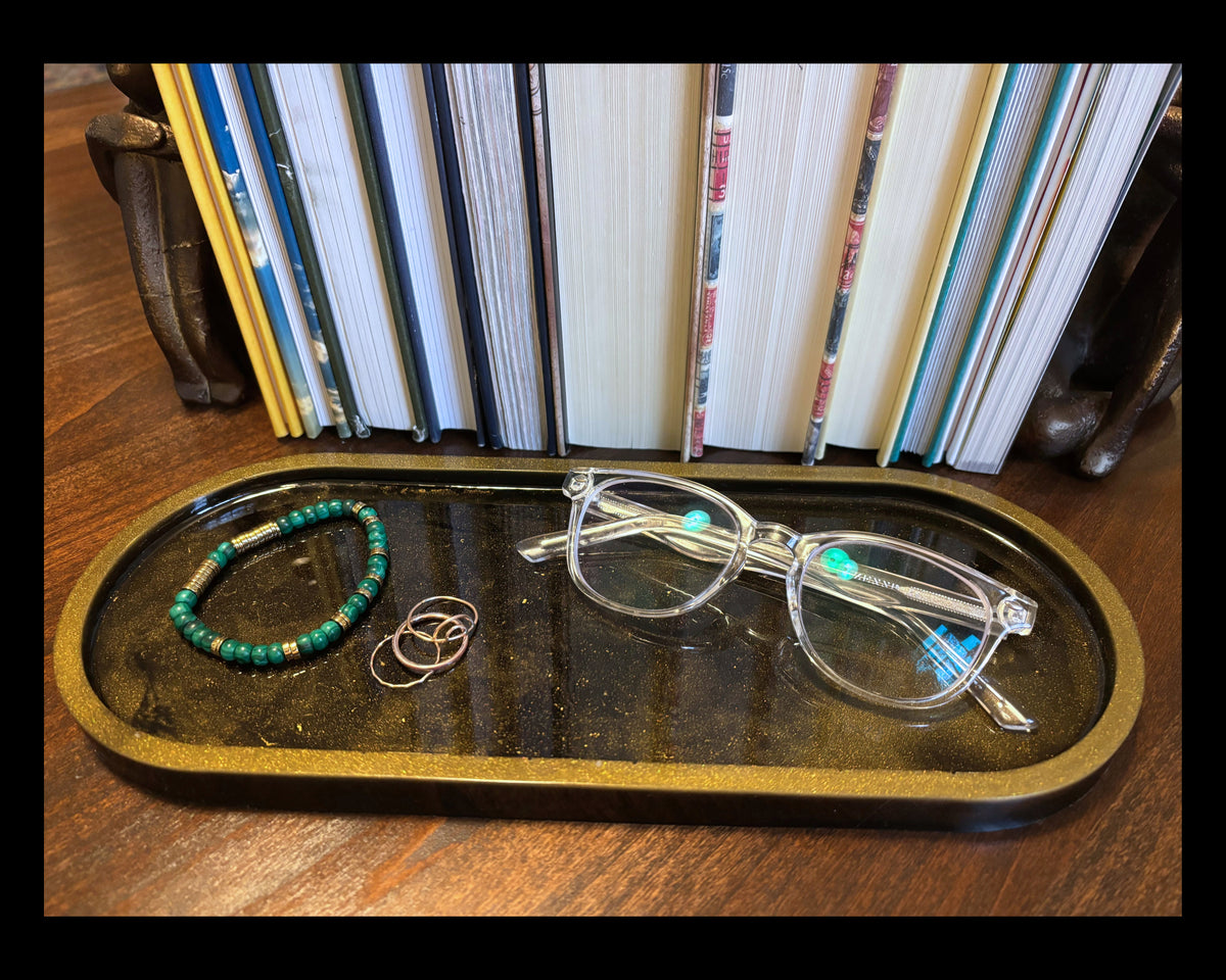 A pair of clear-framed eyeglasses  rests on a black oval tray with a gold-toned rim, alongside a green beaded bracelet and two rings, set against a background of upright books.