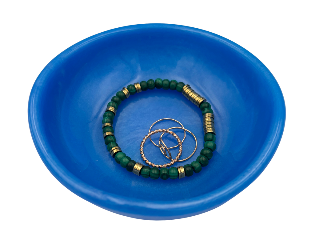 A blue resin bowl containing a green beaded bracelet with gold spacer beads and three gold rings, photographed on a solid white background.