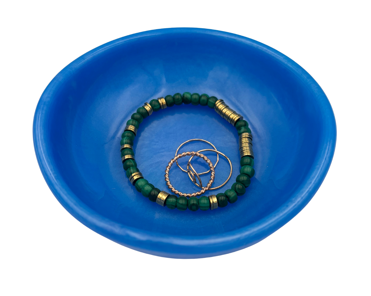 A blue resin bowl containing a green beaded bracelet with gold spacer beads and three gold rings, photographed on a solid white background.