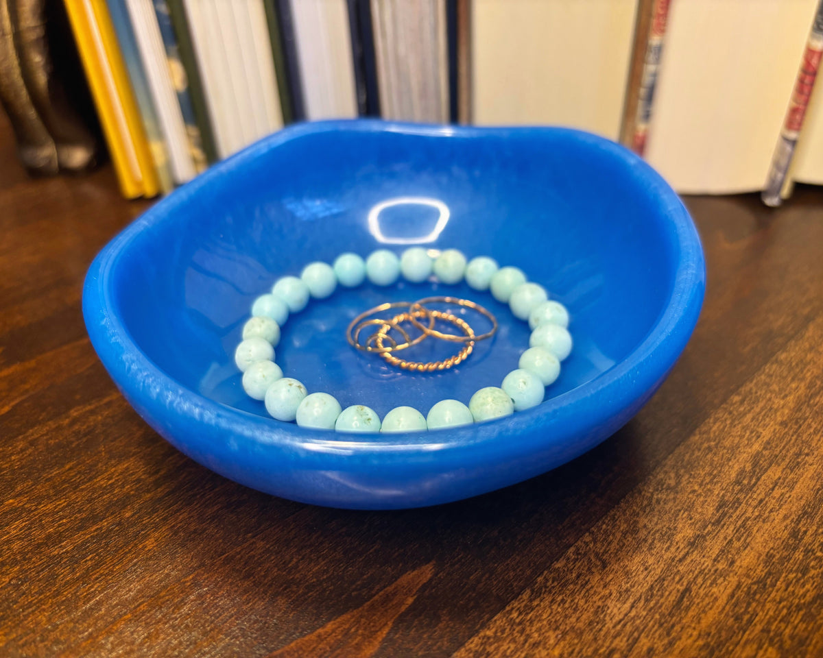 A light blue beaded bracelet with two gold rings resting inside a glossy cobalt blue resin dish on a dark wooden table, with books blurred in the background.