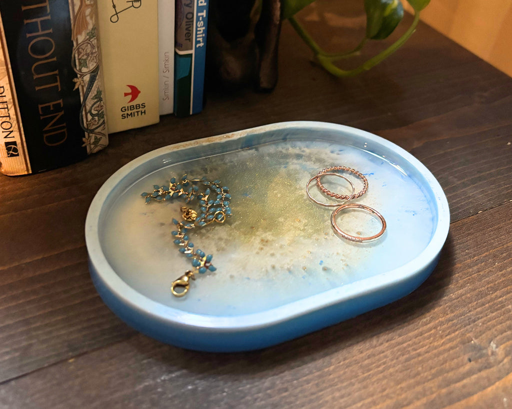 A light blue oval resin jewelry organizer tray with white and gold features, holding jewelry, placed on a dark wooden table next to a bookshelf.