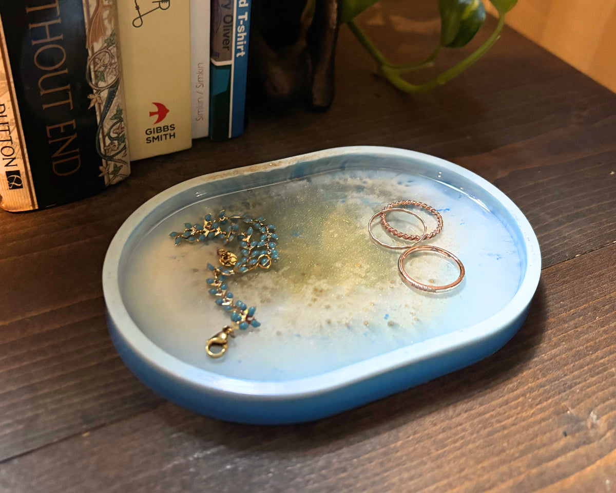 A light blue oval resin jewelry organizer tray with white and gold features, holding jewelry, placed on a dark wooden table next to a bookshelf.