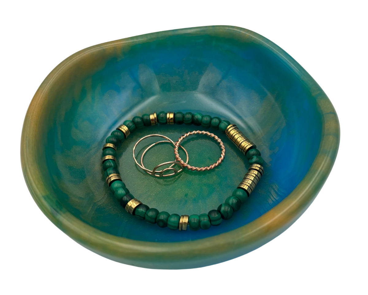 A small organic-shaped resin jewelry tray in a teal and green marbled finish, containing two beaded bracelets and two metal rings, isolated on a white background.