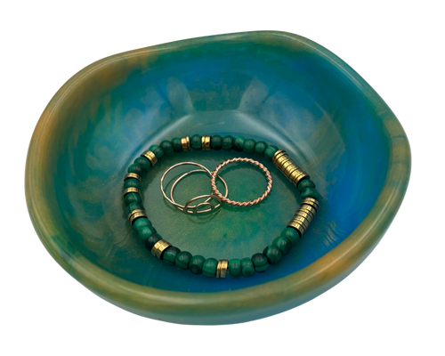 A small organic-shaped resin jewelry tray in a teal and green marbled finish, containing two beaded bracelets and two metal rings, isolated on a white background.