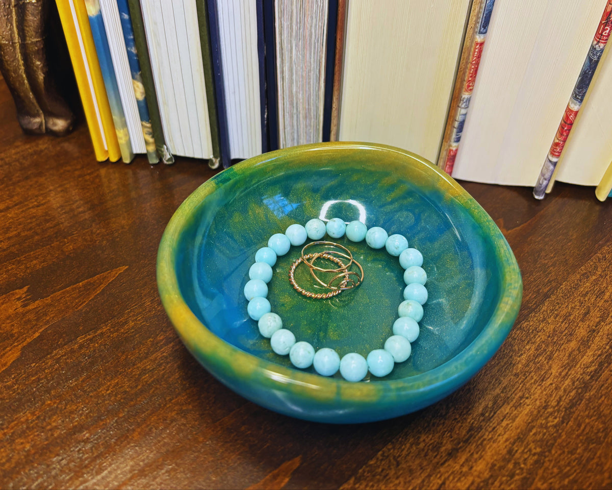 A turquoise and green resin dish on a wooden table, holding a light blue beaded bracelet and two gold rings, with books in the background.