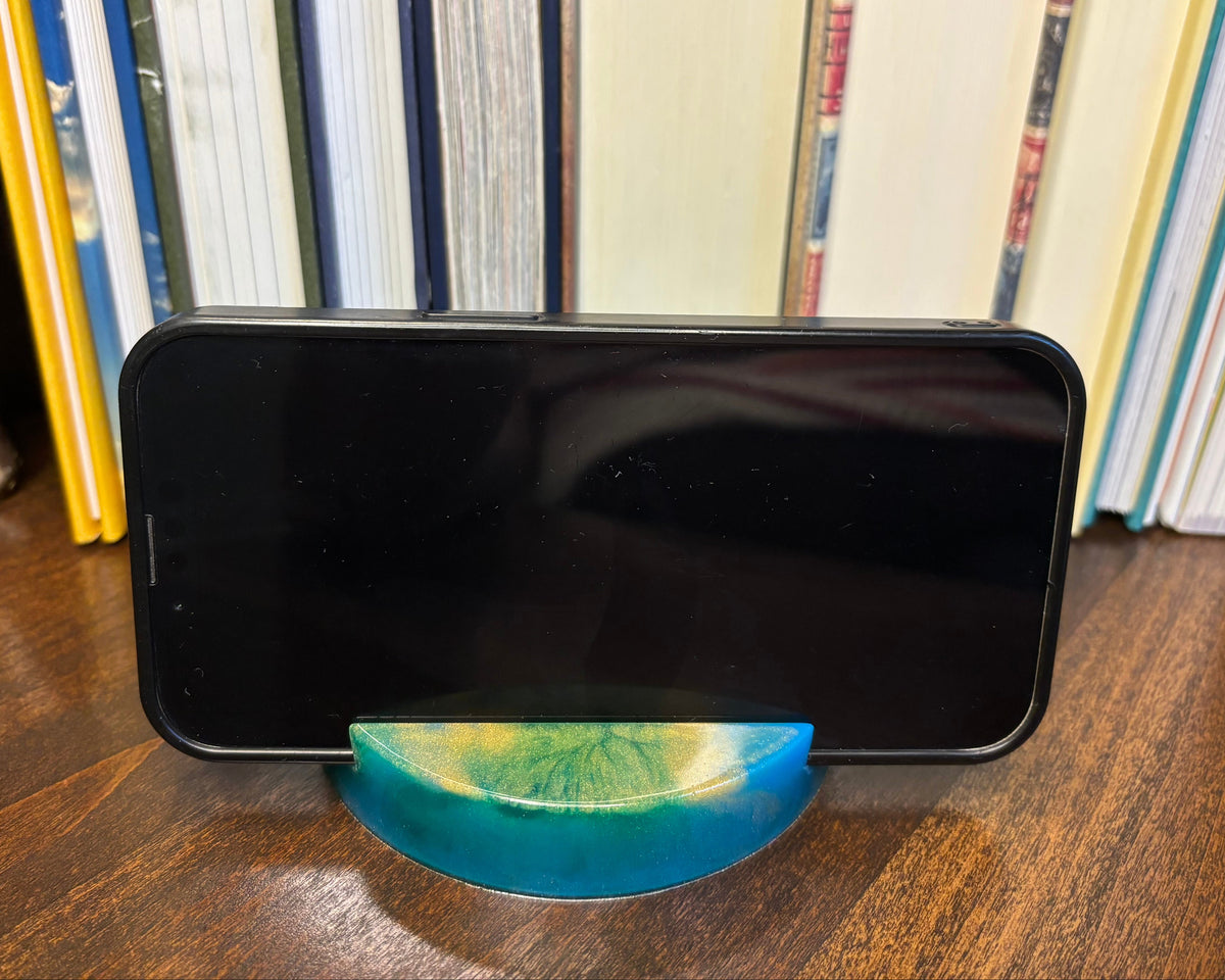 A black smartphone displayed horizontally on a small curved stand made of blue and green marbled resin, set on a wooden surface in front of a bookshelf.