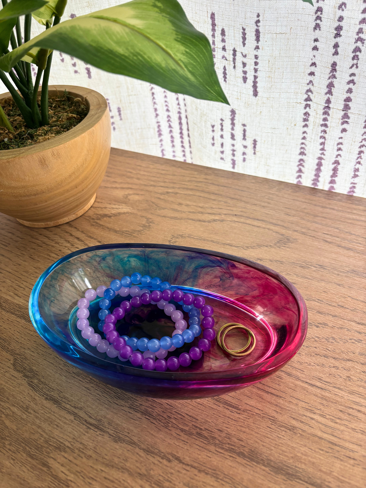 Oval resin dish with blue and pink swirls holding rings and bracelets on a wooden table