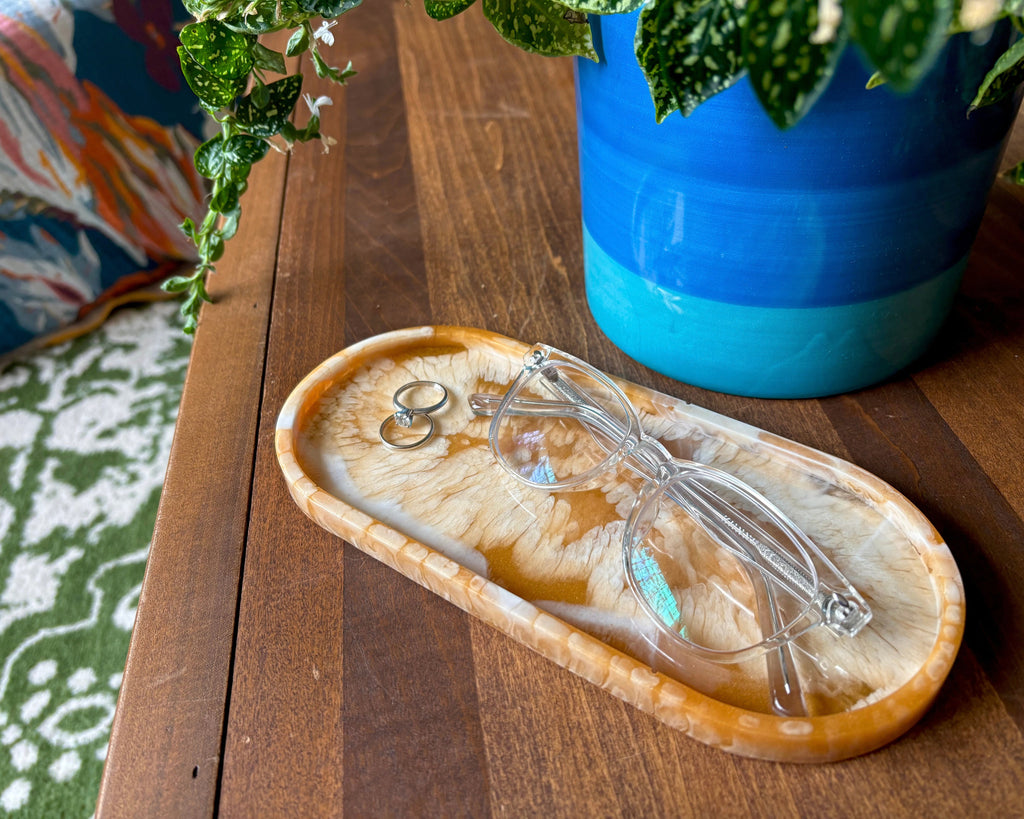 An oval resin tray with a swirling agate-like pattern in shades of brown, tan, and cream, holding glasses and rings on a wooden table
