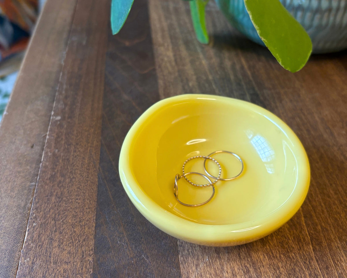 A small round yellow resin dish holding three gold rings, placed on a wooden table with green leaves and a textured vase in the background.