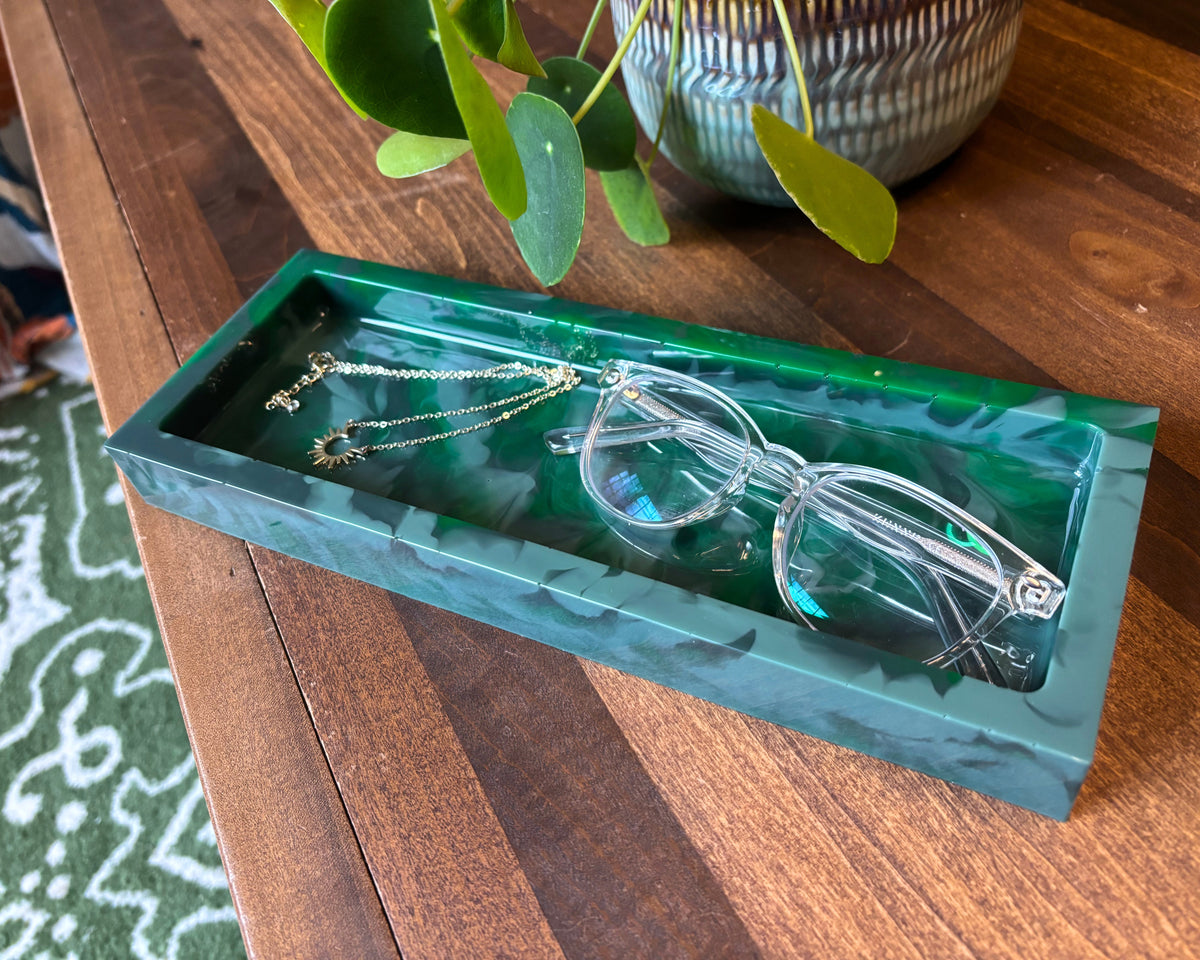 A rectangular green resin tray with a marbled pattern, styled on a wooden table with eyeglasses and a gold necklace inside.