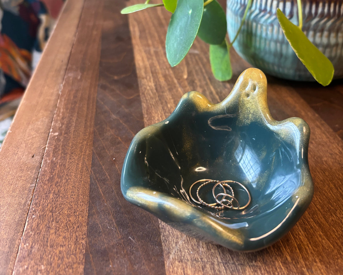 A small decorative bowl with a wavy, ghost silhouette, featuring a glossy dark green to black marbled finish with prominent golden-yellow veining and highlights, sitting on a wooden table with a plant