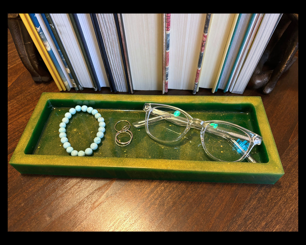 A green rectangular jewelry organizer tray on a wooden table, holding a light blue beaded bracelet, two small rings, and a pair of clear eyeglasses, with a row of books in the background.