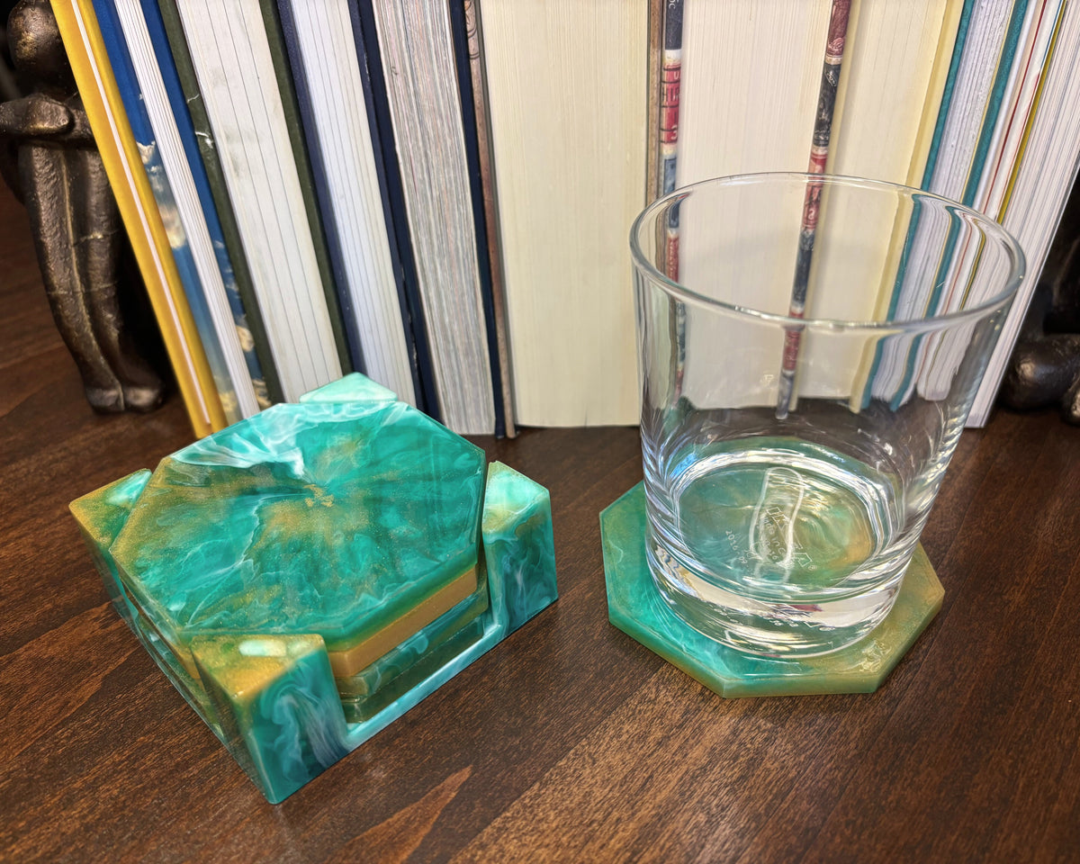 A set of two octagonal resin coasters and matching holder in a green and teal marbled pattern, shown with a clear glass tumbler on a wooden table in front of books.