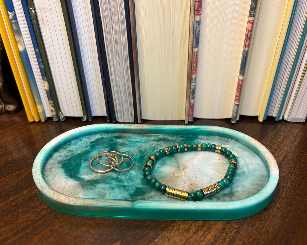 A beaded bracelet with green and gold beads rests on a teal marbled oval tray, which also holds two gold rings, set against a background of standing books.