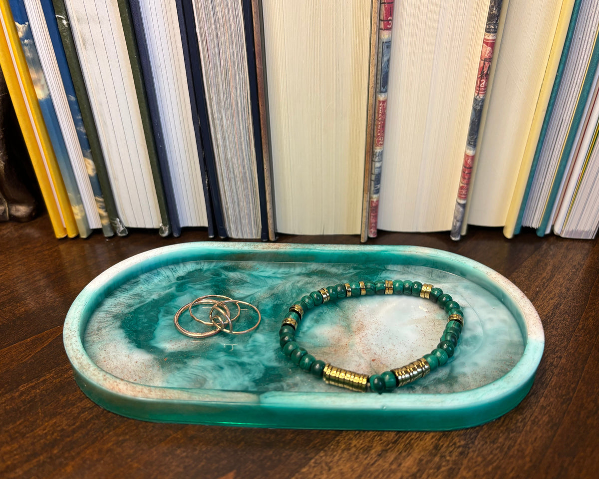 A beaded bracelet with green and gold beads rests on a teal marbled oval tray, which also holds two gold rings, set against a background of standing books.