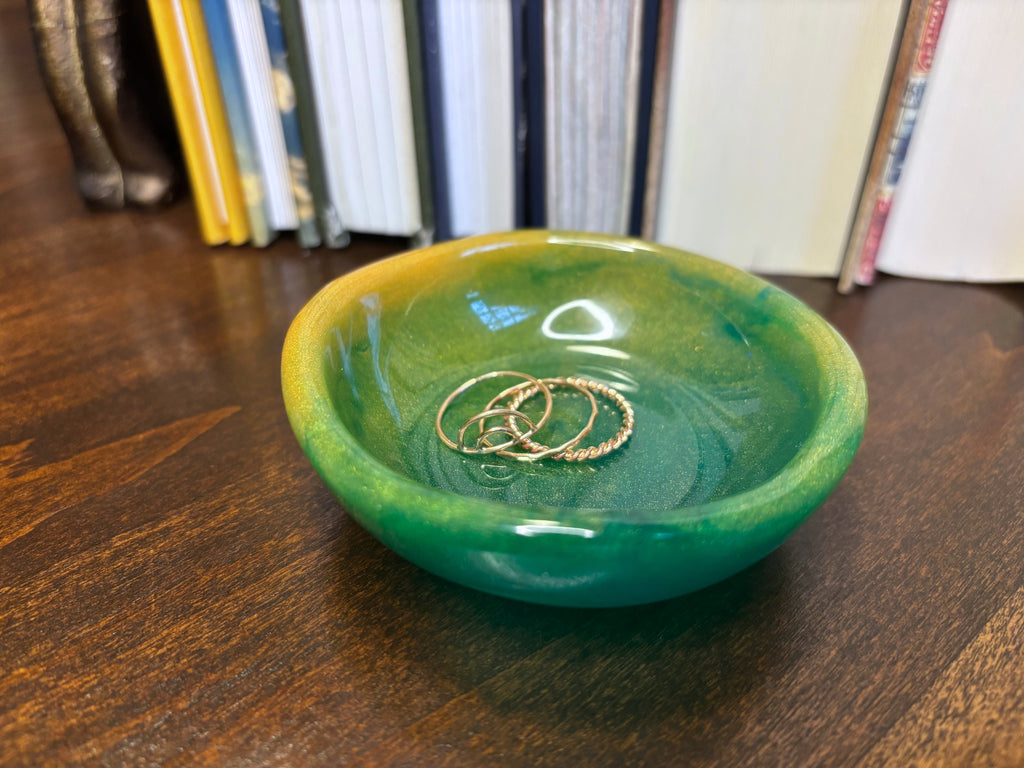 A small round resin jewelry tray with a green and yellow marbled glaze, sitting on a dark wooden table with gold rings inside, and blurred books in the background.