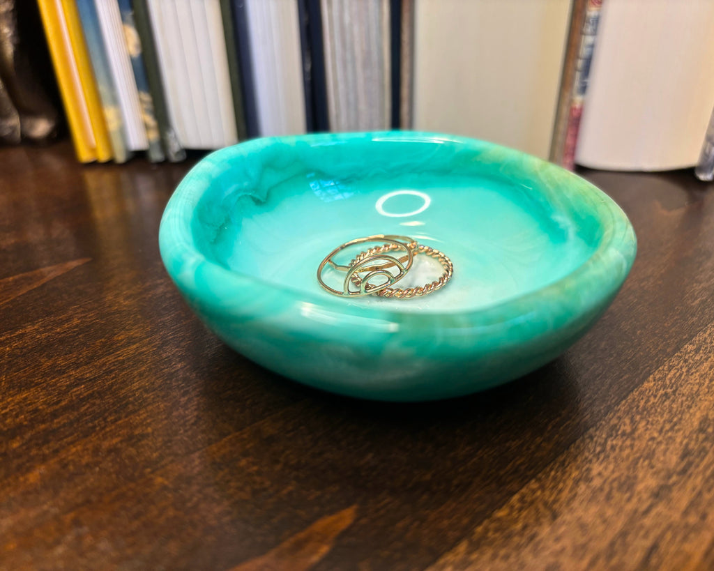 A small round turquoise resin jewelry tray with a marbled pattern, sitting on a dark wooden table with books blurred in the background. Two gold rings are placed inside the tray.