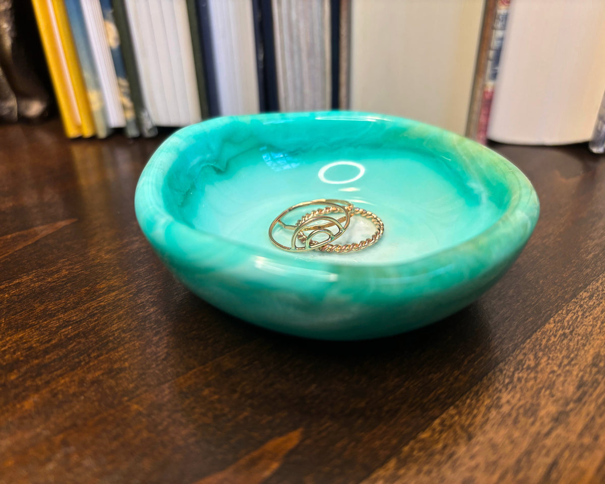 A small round turquoise resin jewelry tray with a marbled pattern, sitting on a dark wooden table with books blurred in the background. Two gold rings are placed inside the tray.