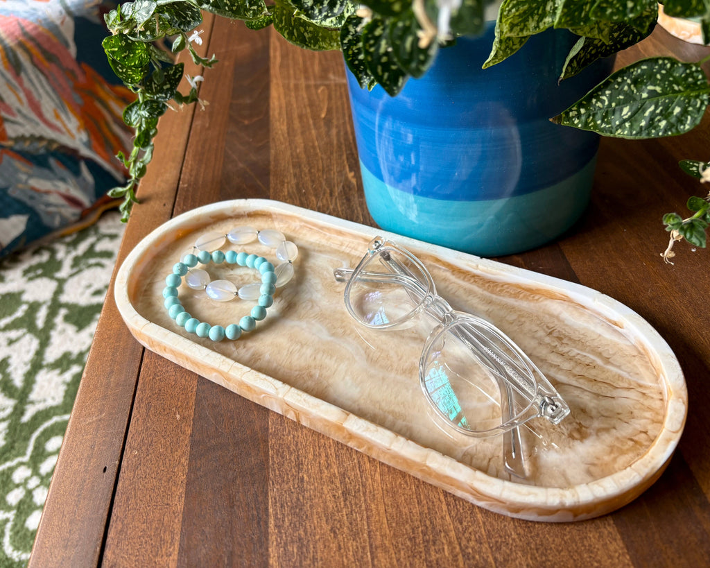 A clear-framed pair of eyeglasses rests on a light resin oval tray alongside a turquoise beaded bracelet, placed on a wooden table with a blue ceramic planter and green leaves in the background.