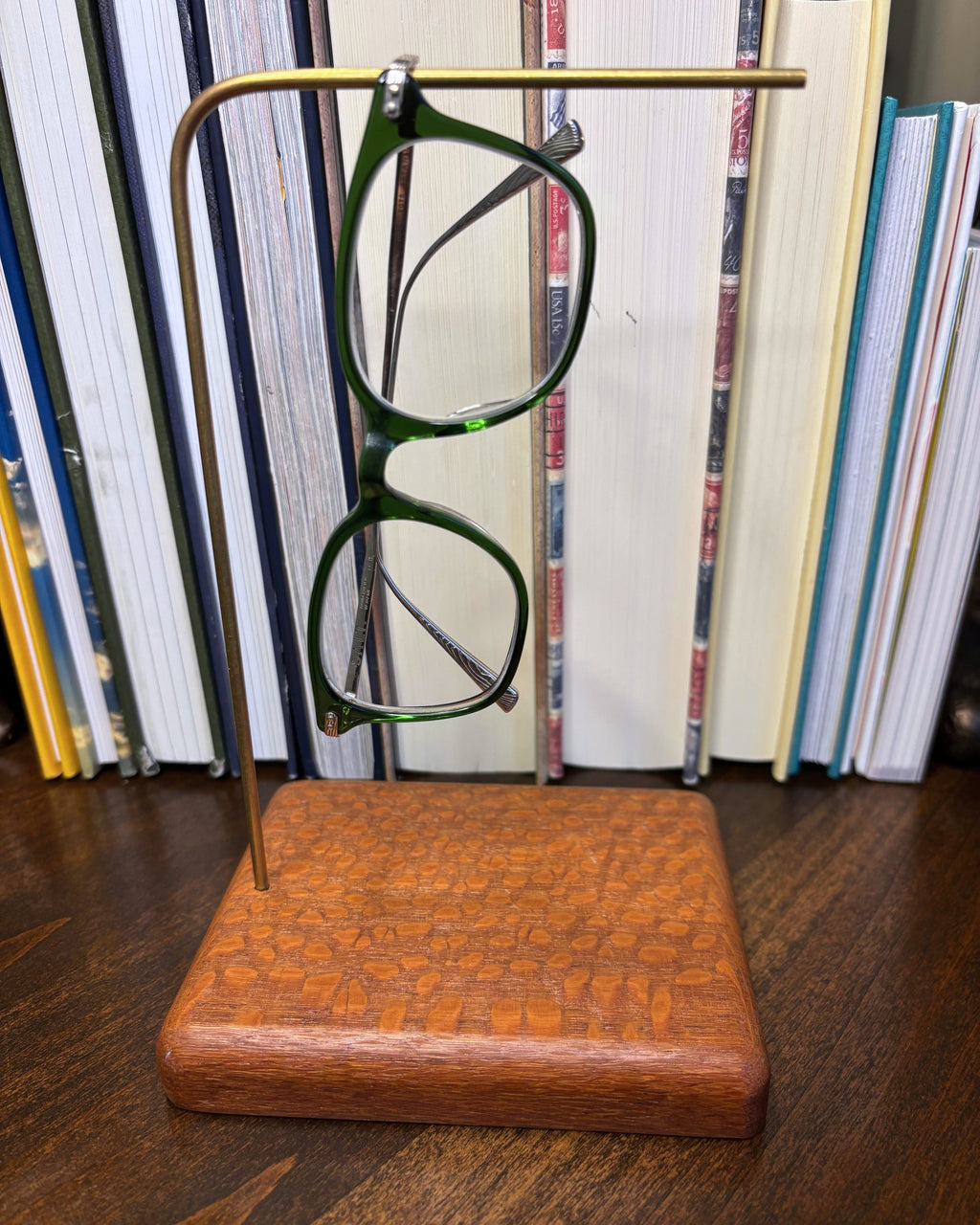 Green eyeglasses displayed on a brass and wood stand in front of a bookshelf.