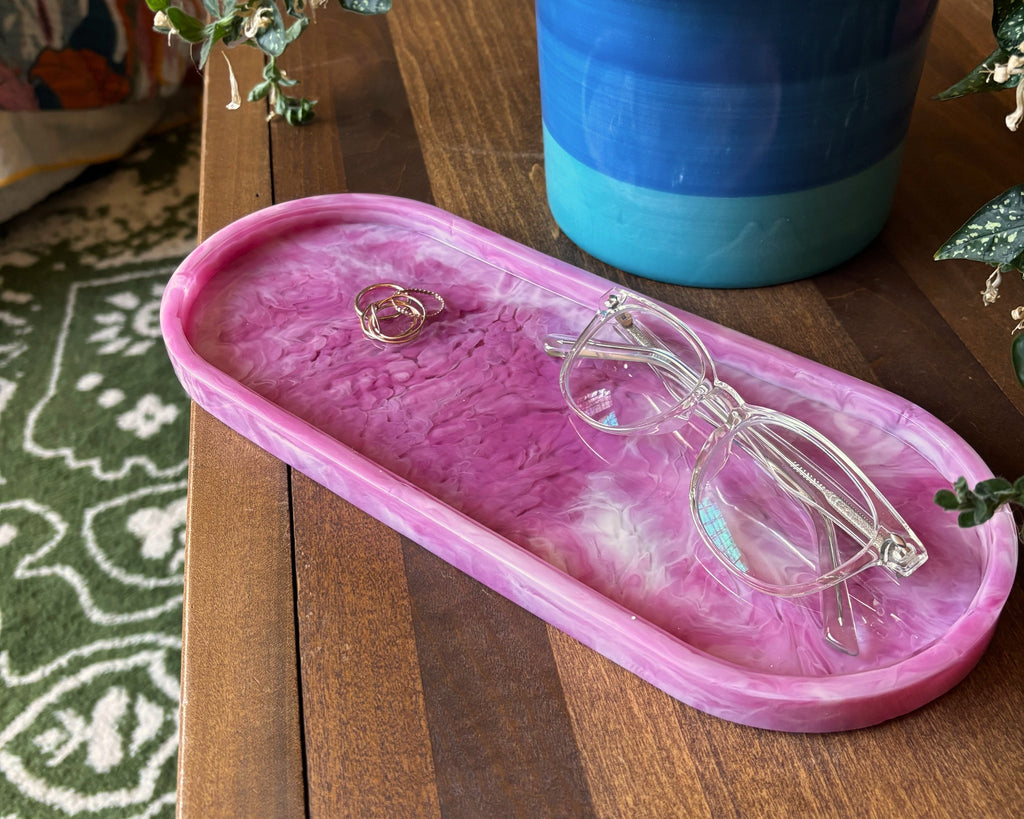 A pink marbled resin jewelry tray with a feather accent, styled on a wooden table next to a potted plant with glasses resting on it.