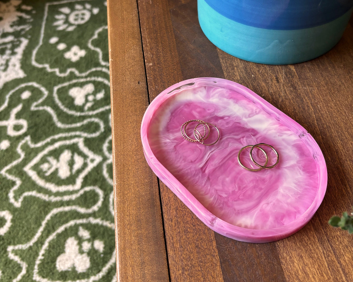 A pink and white marbled oval ring tray holding two gold rings, placed on a wooden table next to a blue and green ceramic planter.