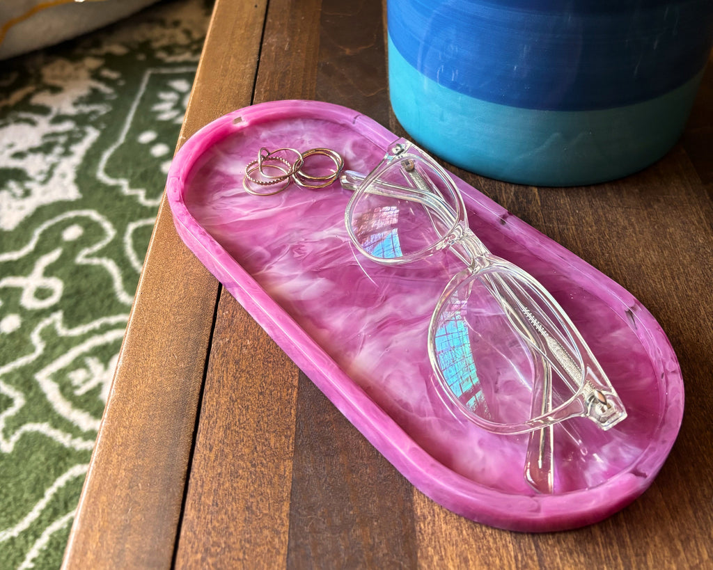 A pink marbled resin eyeglasses tray holding a pair of clear eyeglasses, placed on a wooden table next to a blue and green ceramic planter.