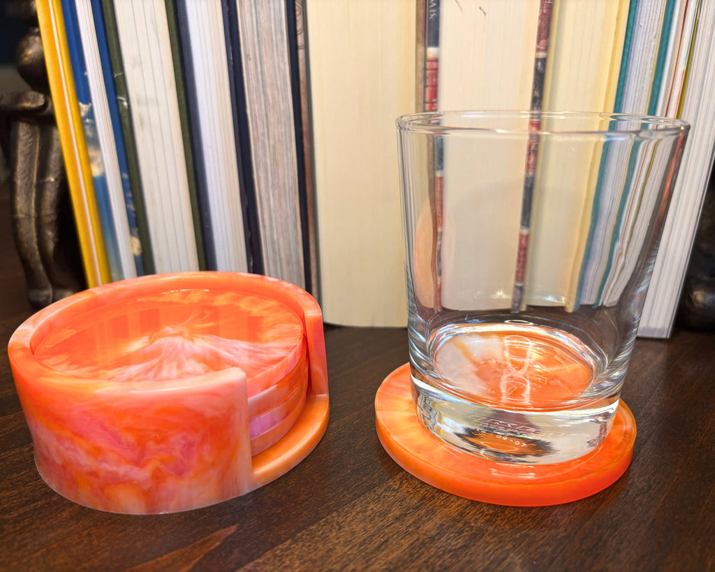An orange and pink marbled resin coaster set on a dark wooden table, featuring a round coaster holding a clear glass tumbler.