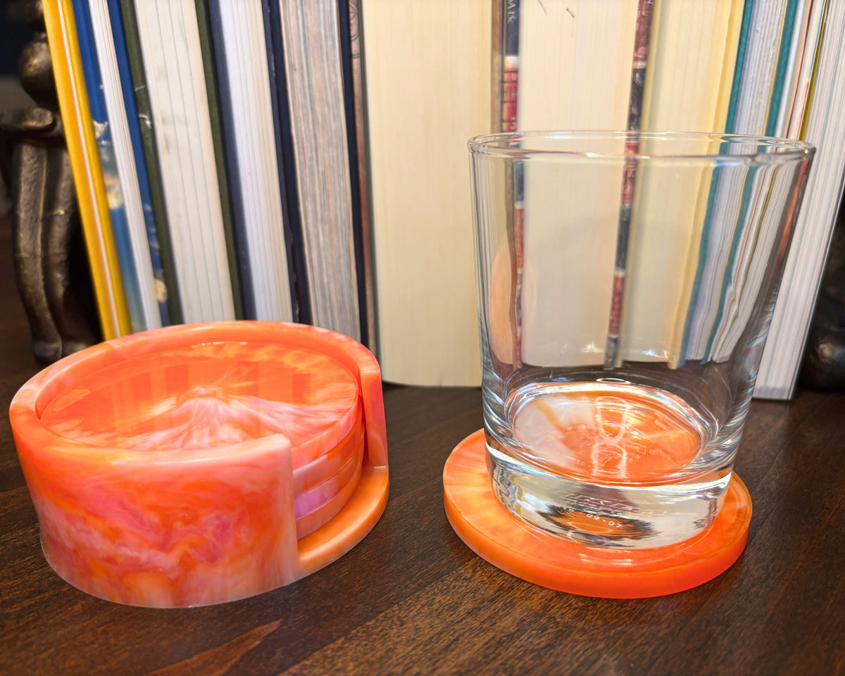 An orange and pink marbled resin coaster set on a dark wooden table, featuring a round coaster holding a clear glass tumbler.