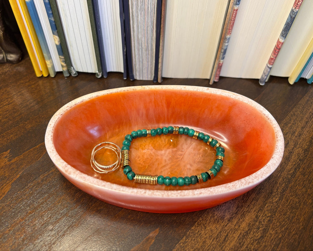 A green beaded bracelet and two gold rings displayed inside a small oval resin dish with a terracotta-orange interior, set on a dark wooden tabletop with books in the background.