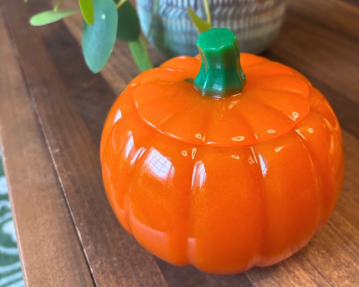 An orange pumpkin-shaped resin container with a green stem lid, sitting on a wooden table next to a potted plant.