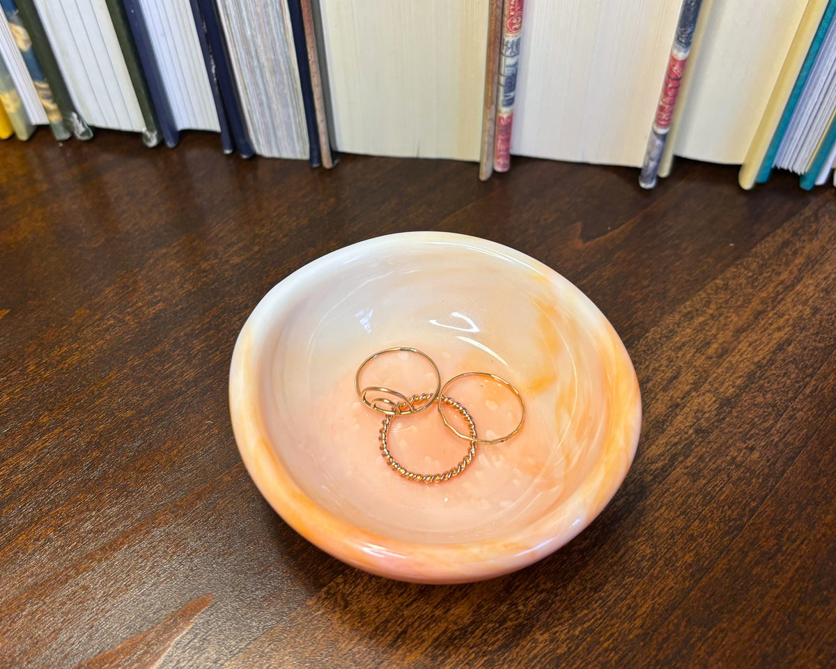 A small round marbled bowl in peach, pink, and cream sits on a dark wood table, holding three gold rings, with a row of books in the background.