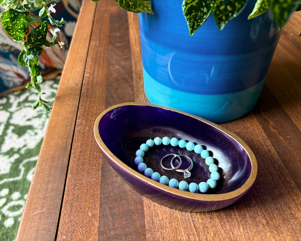 A small oval purple resin dish with a gold-toned rim sits on a wooden table, holding a light blue beaded bracelet and two silver rings. A blue ceramic planter and green leaves are in the background.