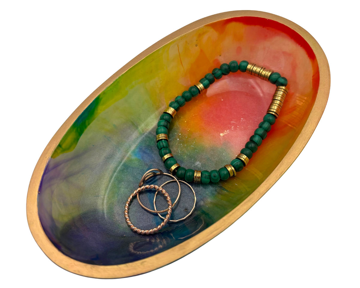 A green beaded bracelet with gold spacer beads and three textured gold rings, displayed on a colorful oval tray with a gold rim.