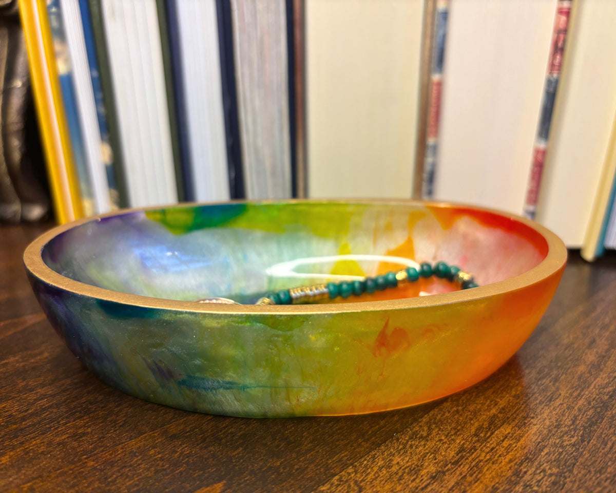 A shallow round bowl with a marbled rainbow finish in green, yellow, orange, and blue, sitting on a dark wooden table with a beaded necklace inside.