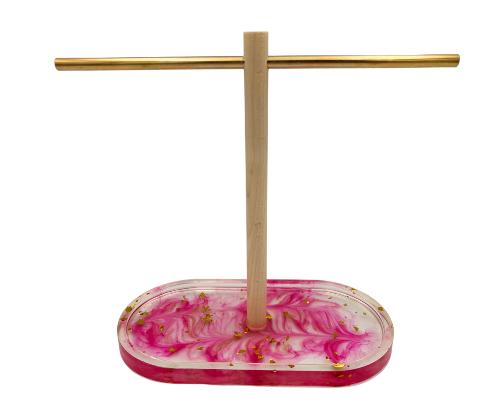 A jewelry display stand with a rectangular pink and white marbled resin base containing small gold flakes, a vertical wooden rod, and a horizontal gold metal bar, shown on a plain white background.