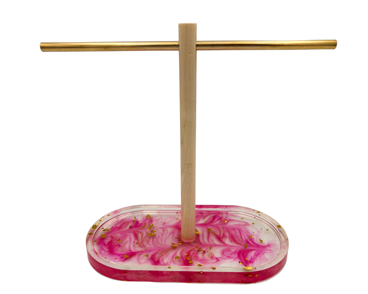 A jewelry display stand with a rectangular pink and white marbled resin base containing small gold flakes, a vertical wooden rod, and a horizontal gold metal bar, shown on a plain white background.