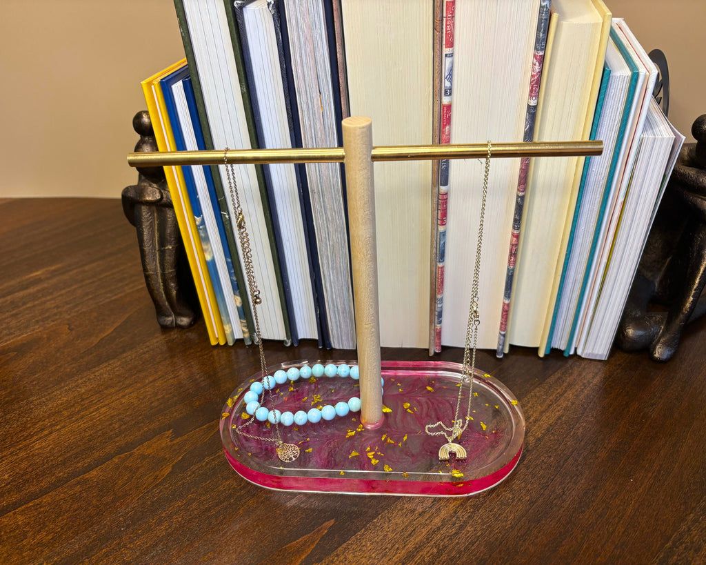 A jewelry display stand with a gold horizontal bar, a light wood central post, and a clear acrylic base with pink glitter. The stand is shown holding a light blue beaded bracelet and silver chain necklaces, set on a dark wooden table in front of a bookshelf.