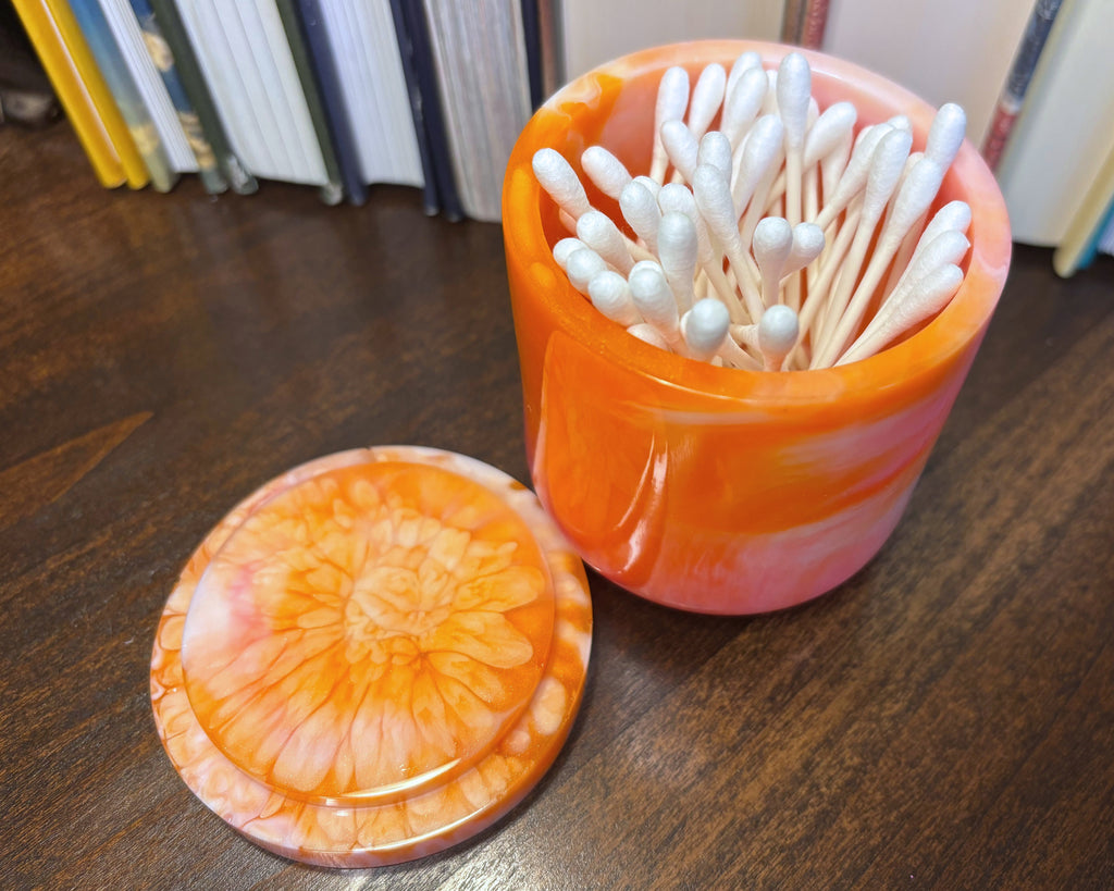 An orange and pink marbled cotton swab container filled with white cotton swabs, shown on a dark wooden table with its matching lid placed in front. Books are visible in the background.