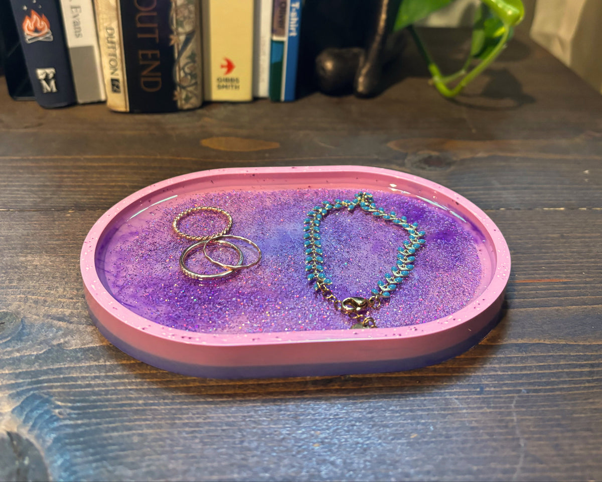 A small oval jewelry tray with a pink rim and a purple glittery interior, holding rings and a beaded bracelet, placed on a dark wooden table with books and a plant in the background.