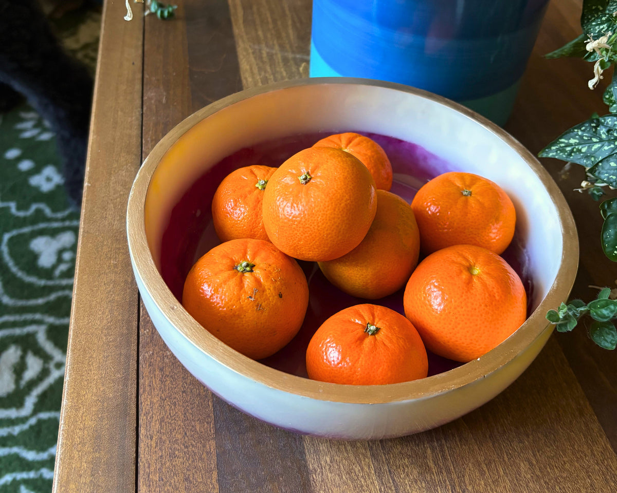 resin bowl in cream and plum tones with a gold rim, holding tangerines, set on a wooden table with a plant in the background