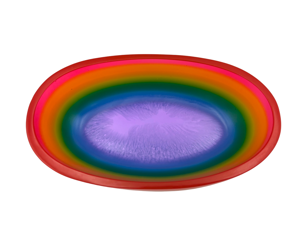 A shallow oval resin bowl with a vibrant rainbow gradient design, featuring concentric bands of red, orange, yellow, green, blue, and purple on a plain white background.