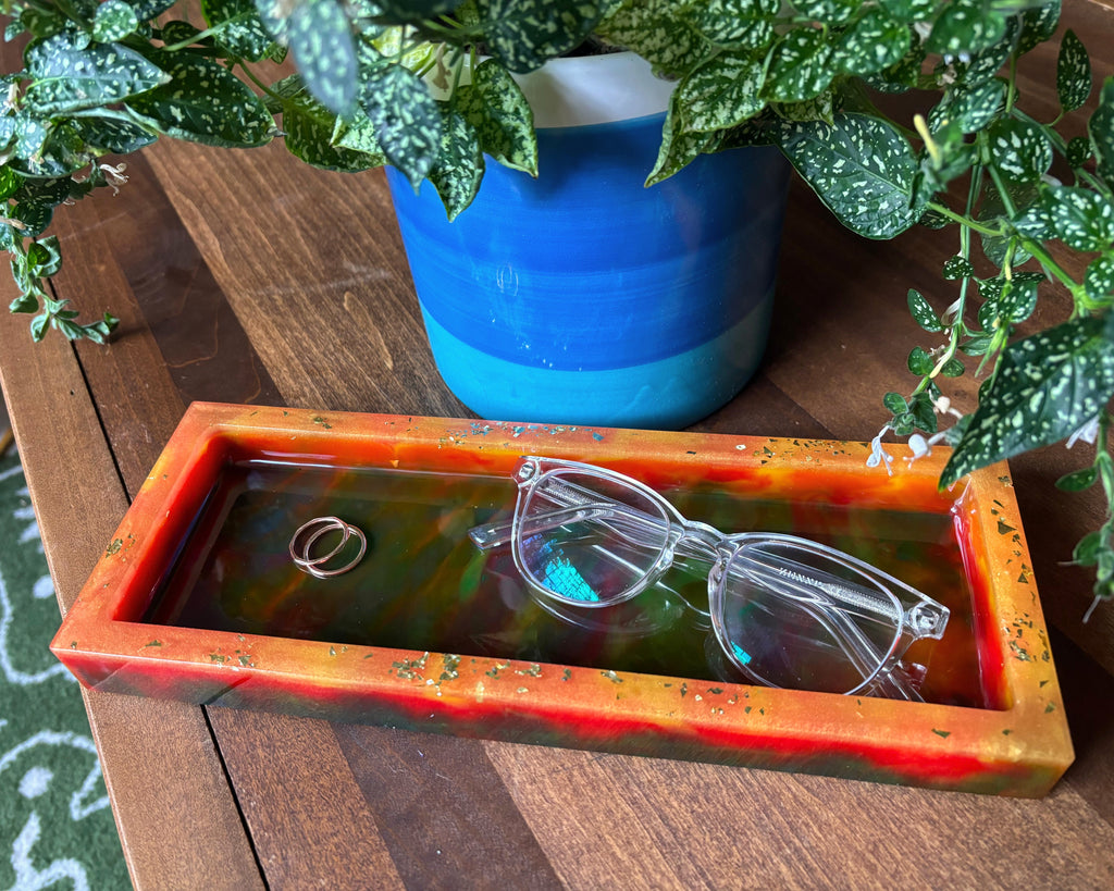 A rectangular resin tray with a marbled orange, red, and green pattern sits on a wooden table. Clear eyeglasses and two rings are placed inside the tray, with a blue and green potted plant in the background.