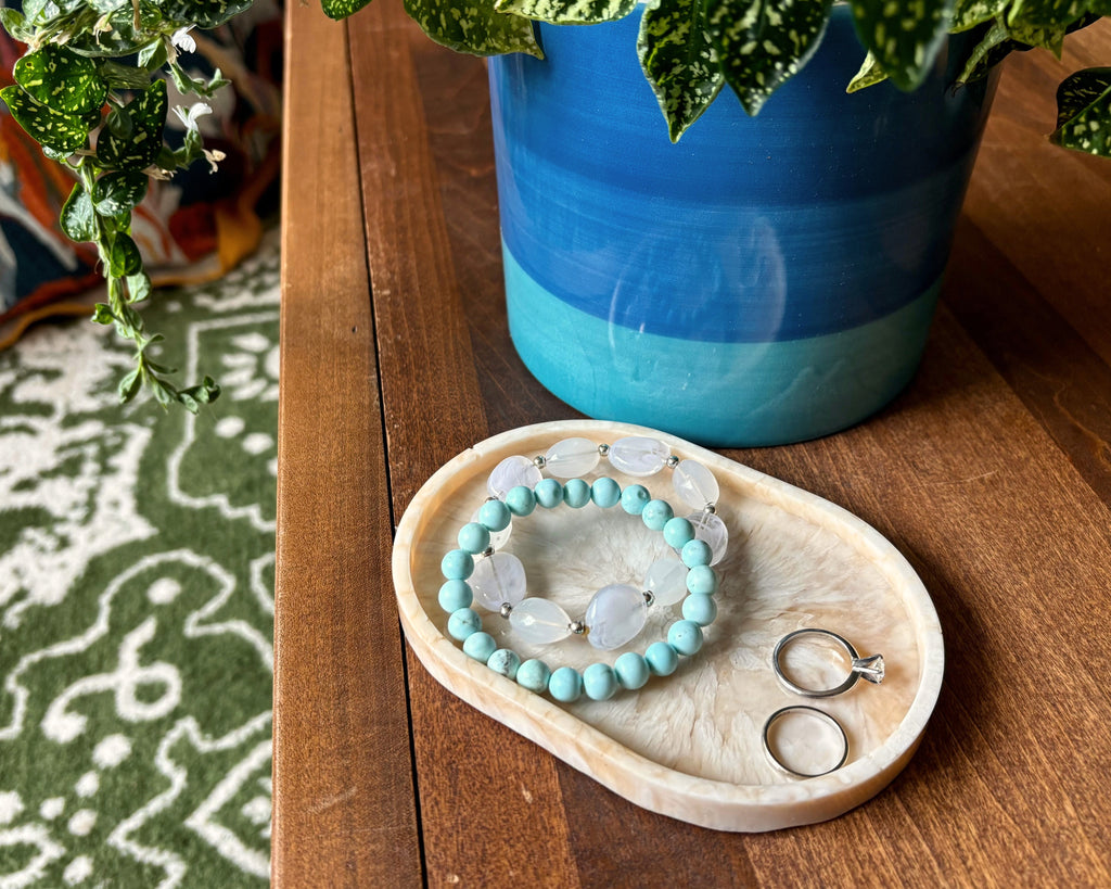 A beaded bracelet with light blue beads and white stone accents, alongside two silver rings, displayed on a small resin tray on a wooden table next to a blue and green ceramic planter.