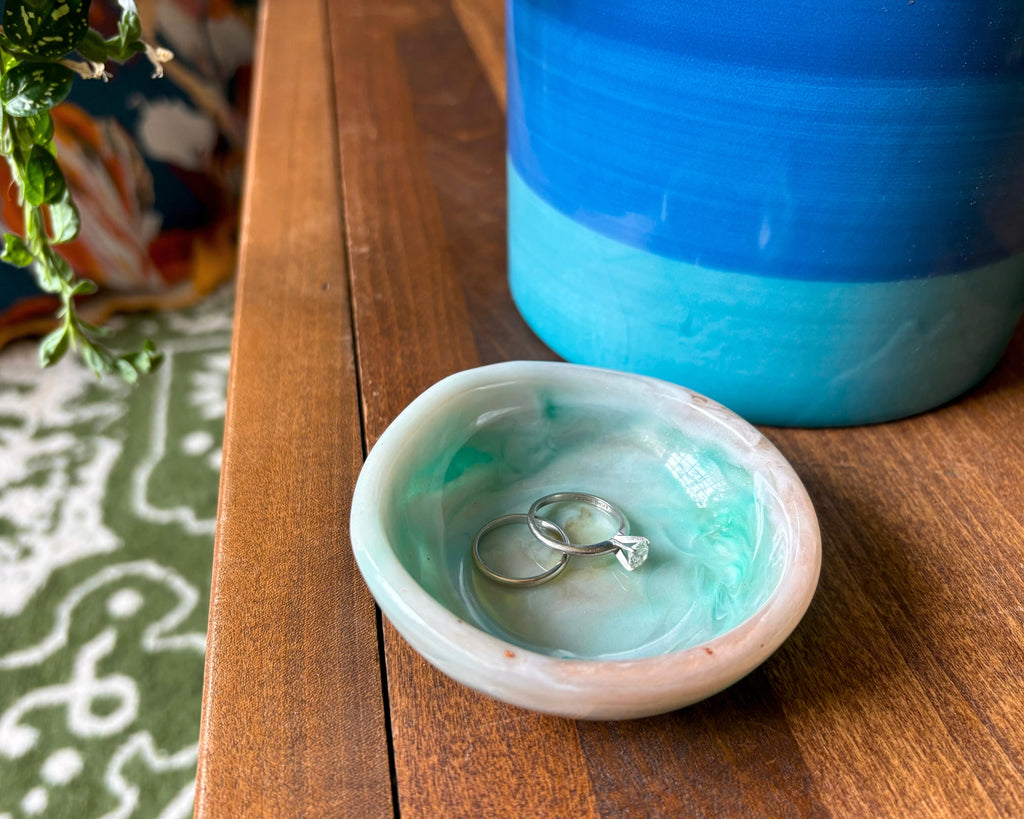 a small ring dish made of resin with teal and beige swirls on a wooden table with rings in the dish.