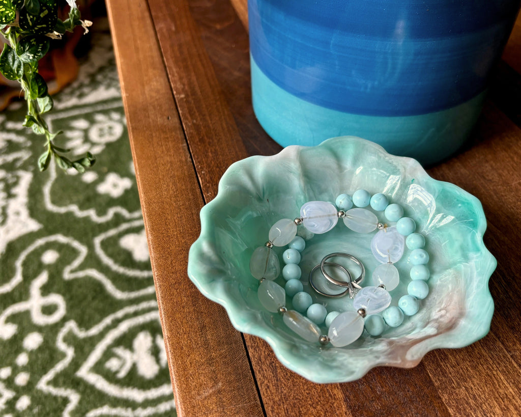 A resin flower-shaped jewelry tray in a light teal and copper pattern, holding a beaded bracelet and two rings, set on a wooden table next to a blue pot.